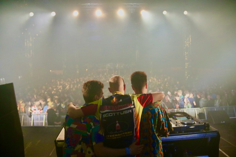 De drie leden van Let's Dance met de armen om elkaar heen, kijkend naar een vol festivalpubliek
