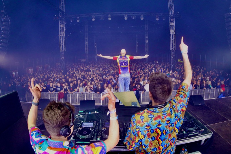 Twee dj's van Let's Dance met de armen in de lucht achter de draaitafels, MC voor een volle festivaltent